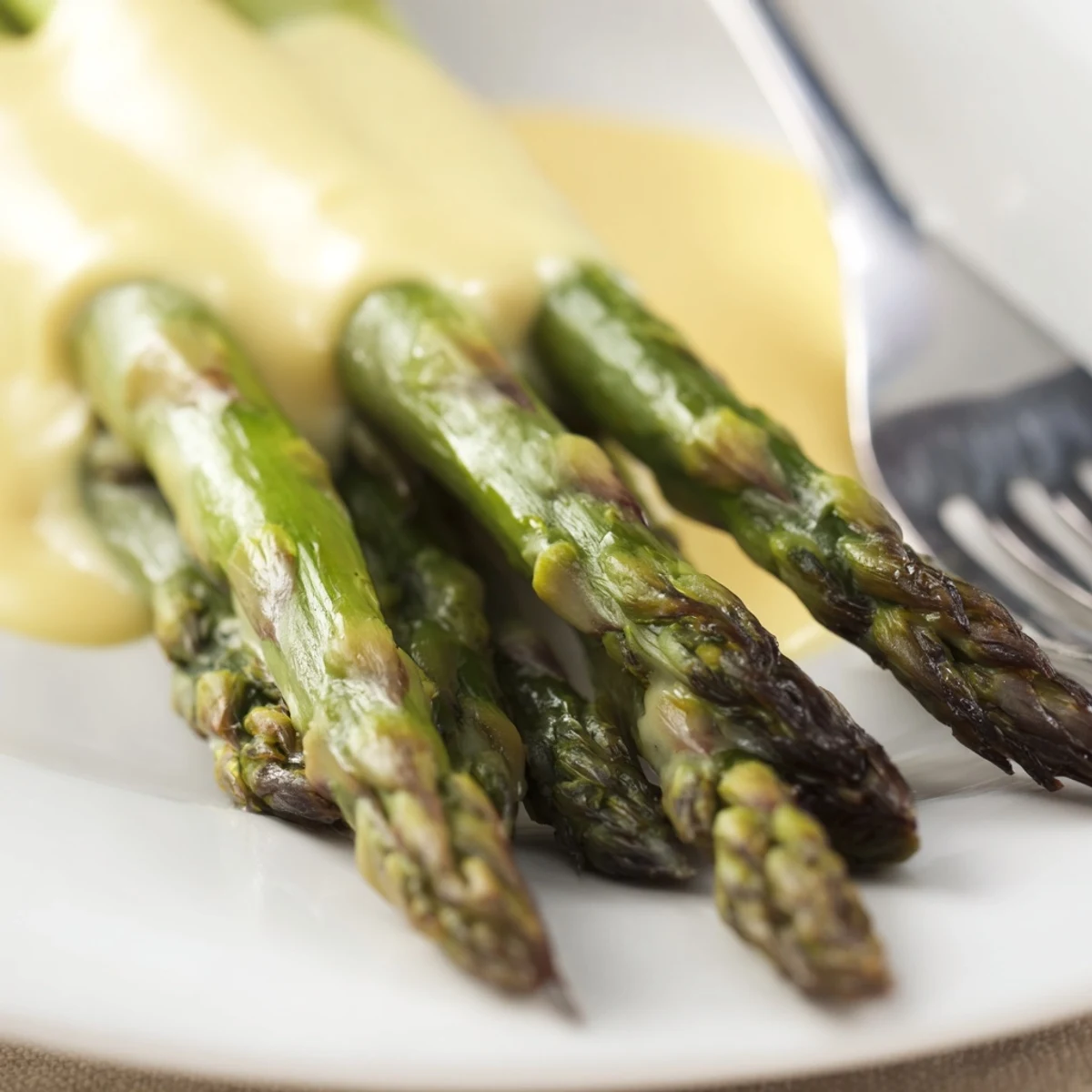 Golden-brown roasted asparagus with Hollandaise sauce glistening on a white platter next to lemon wedges.