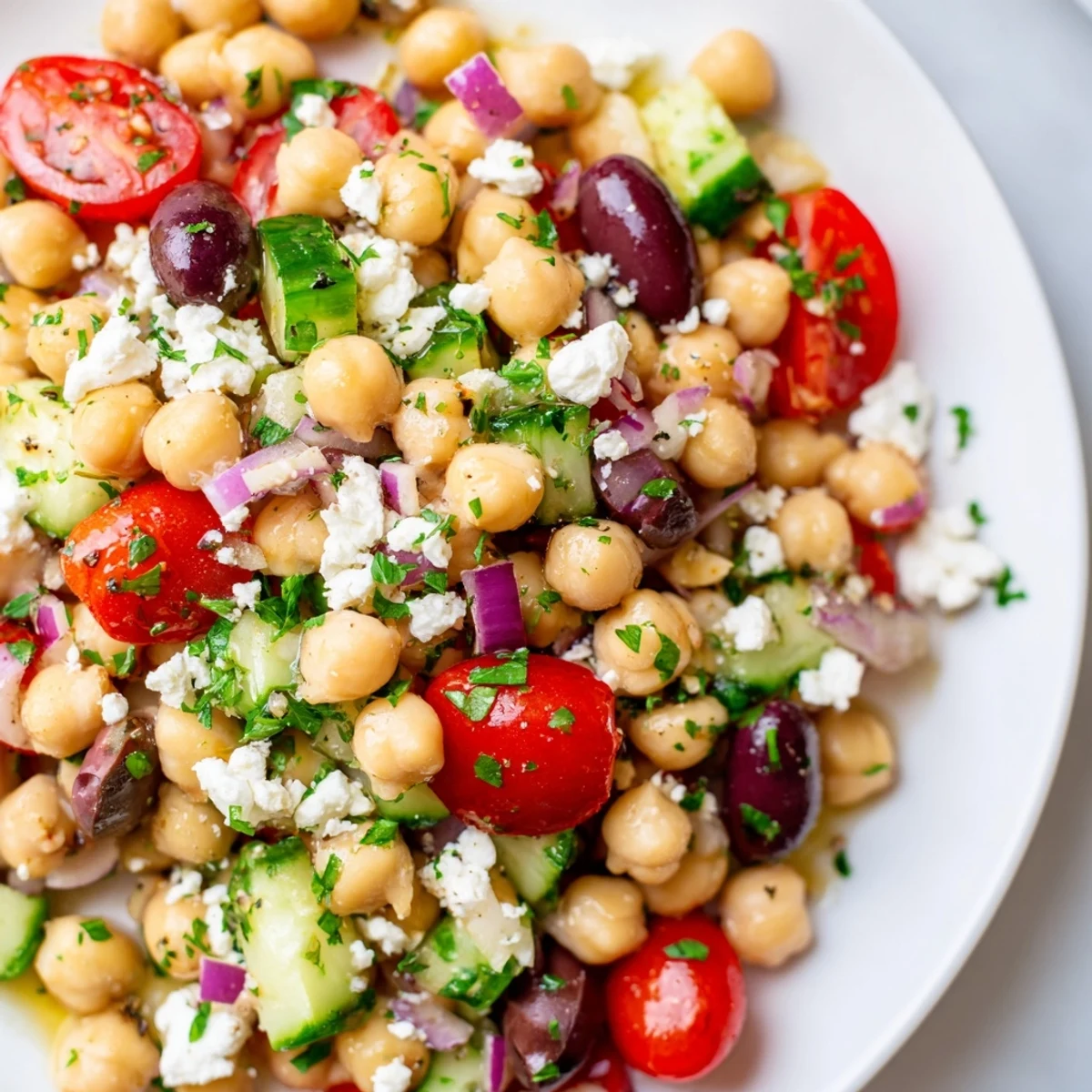 A bowl of Mediterranean Chickpea Salad with Feta shows chopped tomatoes, cucumber, olives, and chickpeas topped with crumbled feta and fresh parsley.