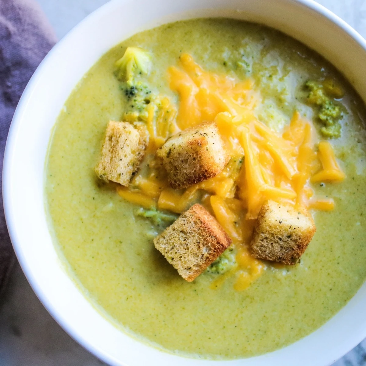 Creamy green broccoli cheddar soup with croutons served hot in a bowl, garnished with extra cheddar and fresh herbs.
