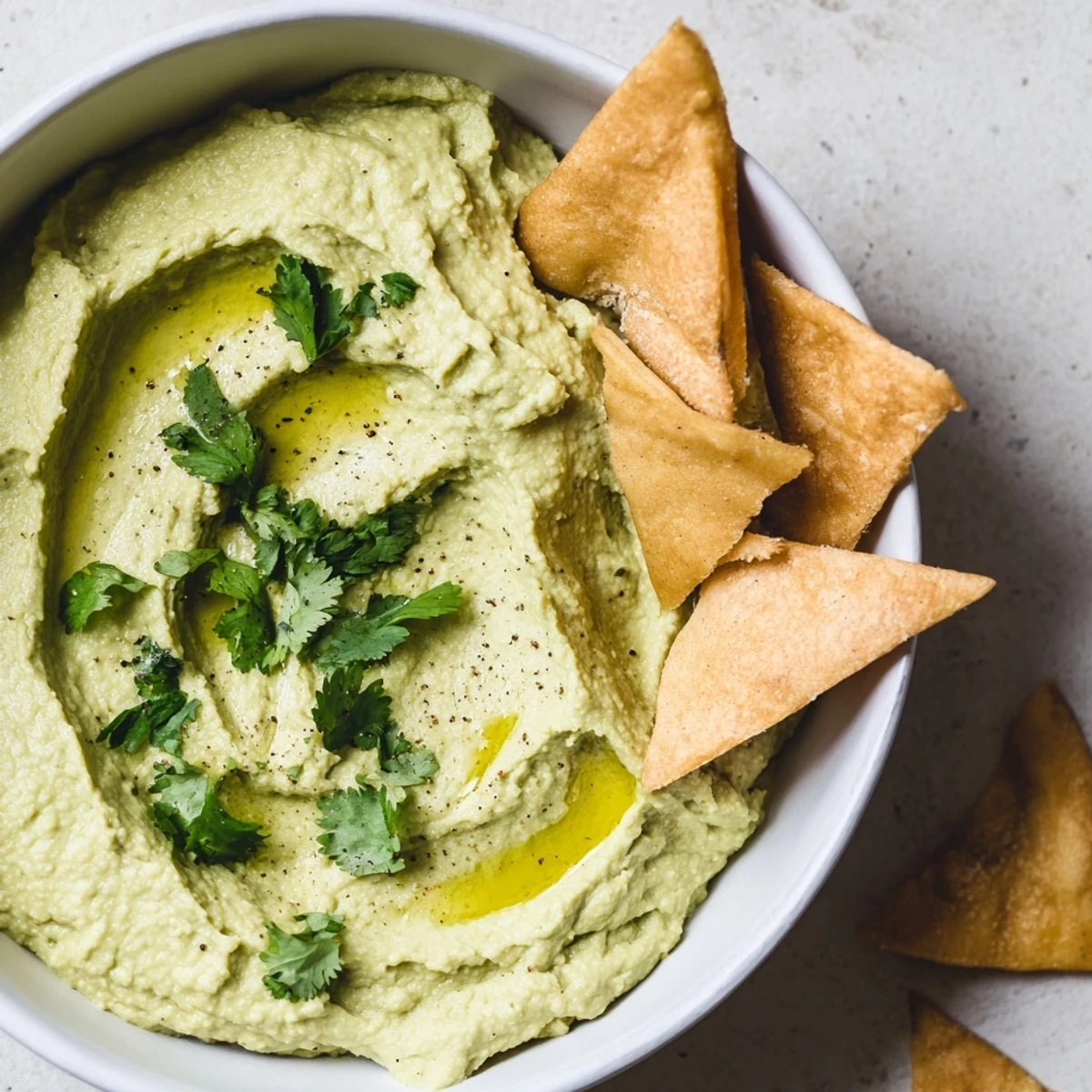 A bowl of creamy Green Avocado Hummus topped with olive oil and herbs, served with golden baked pita chips for dipping.