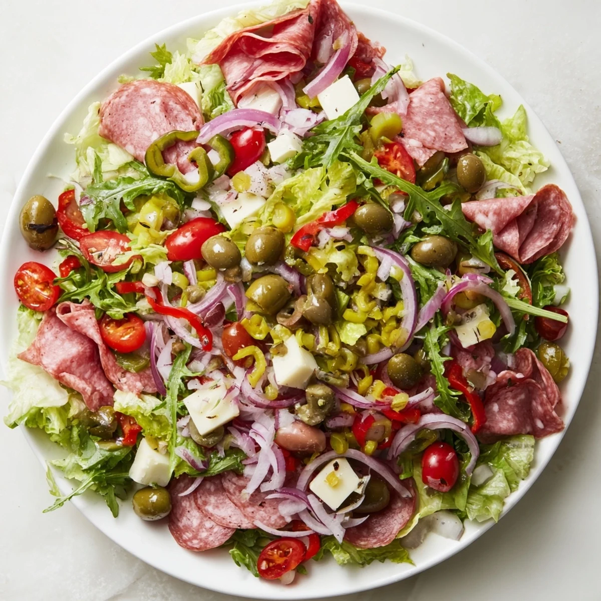 A vibrant New Orleans Muffuletta Salad in a white bowl, featuring chopped romaine, arugula, cherry tomatoes, roasted red peppers, and sliced salami with provolone cubes.