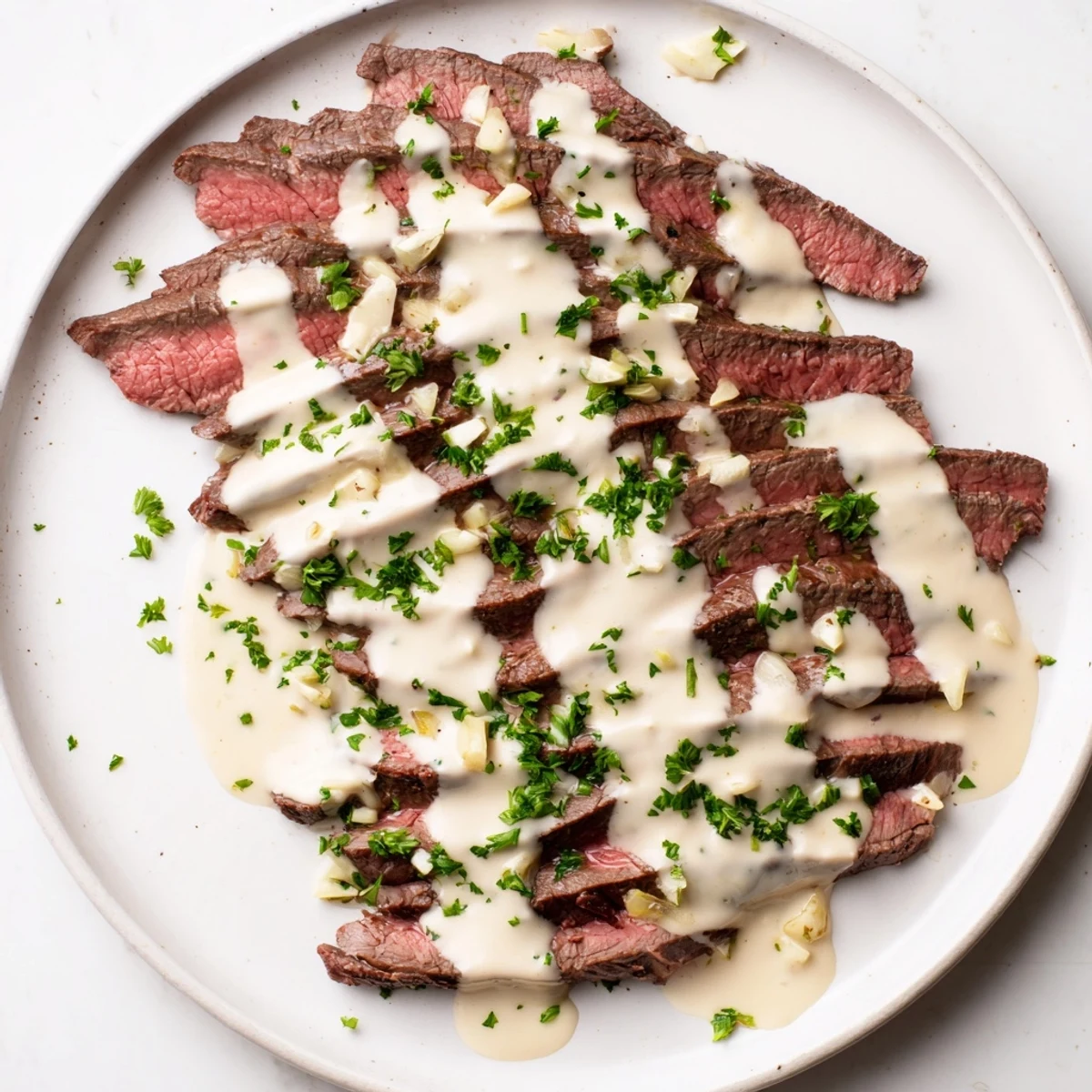 A close-up of flank steak with roasted garlic sauce, showing juicy slices and creamy sauce perfect for serving alongside roasted potatoes or green beans.