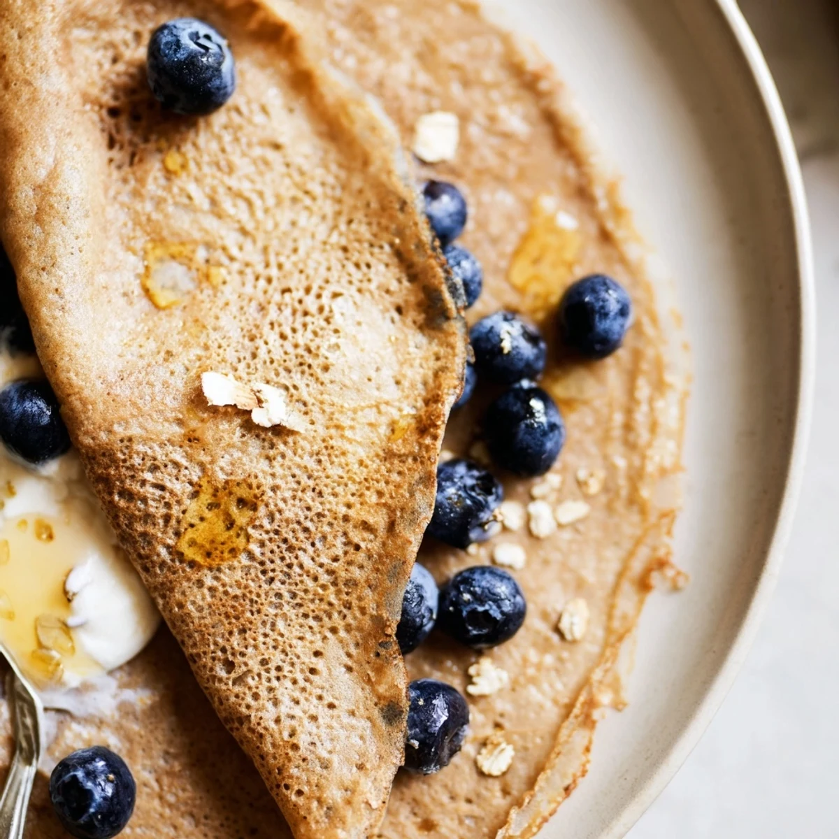 Golden healthy oatmeal crepes stacked on a white plate with fresh berries and honey drizzle