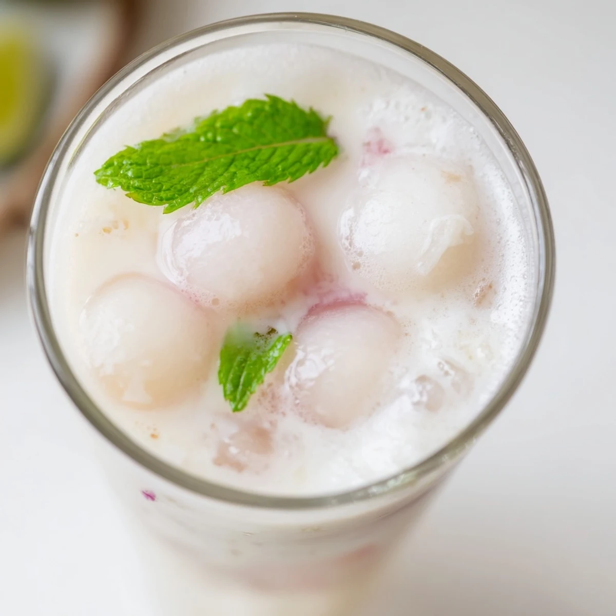 Tall glass of creamy coconut lychee iced drink garnished with fresh mint and lime slices